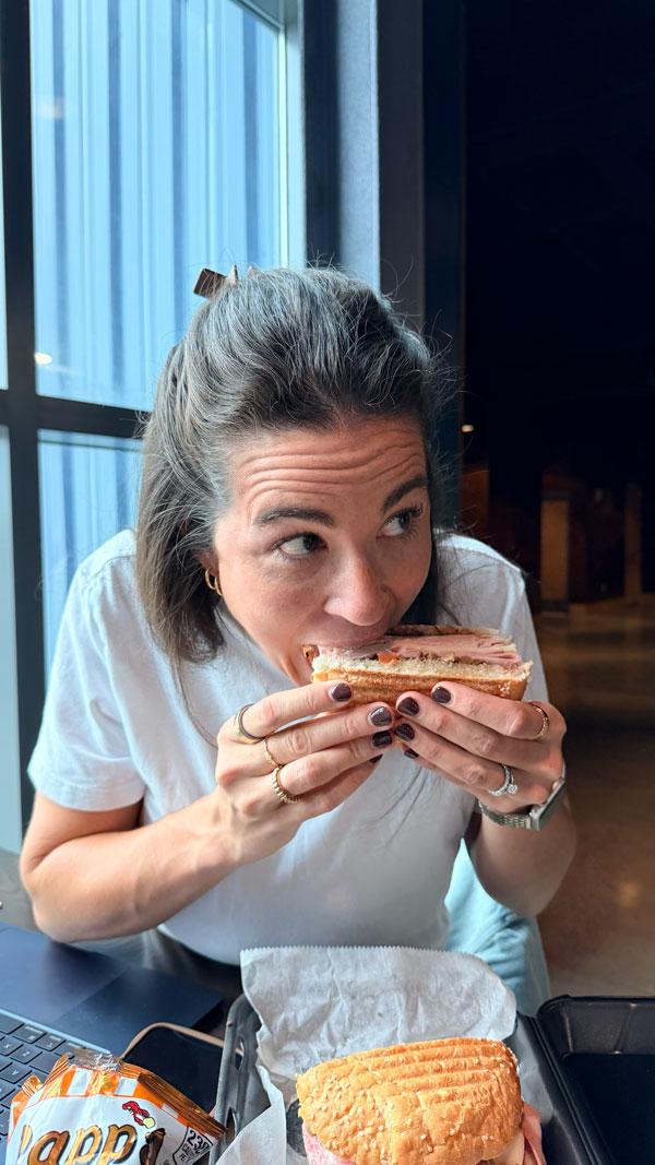 An Ample team member taking a bite of a deli sandwich beside a window