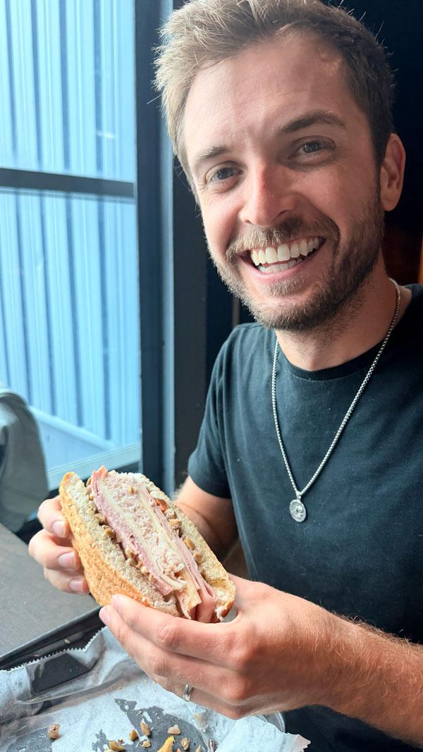 An Ample team member grinning over a deli sandwich beside a window